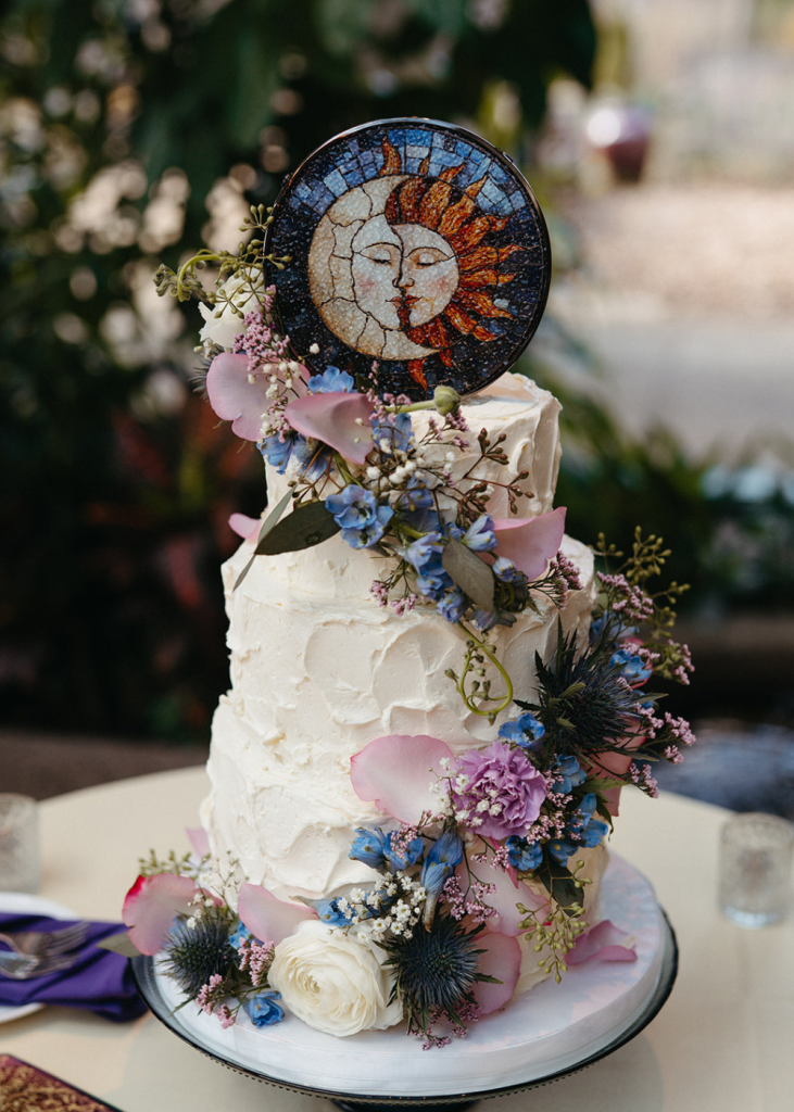 a wedding cake with purple and blue flowers and with a cake topper with glass a moon and sun