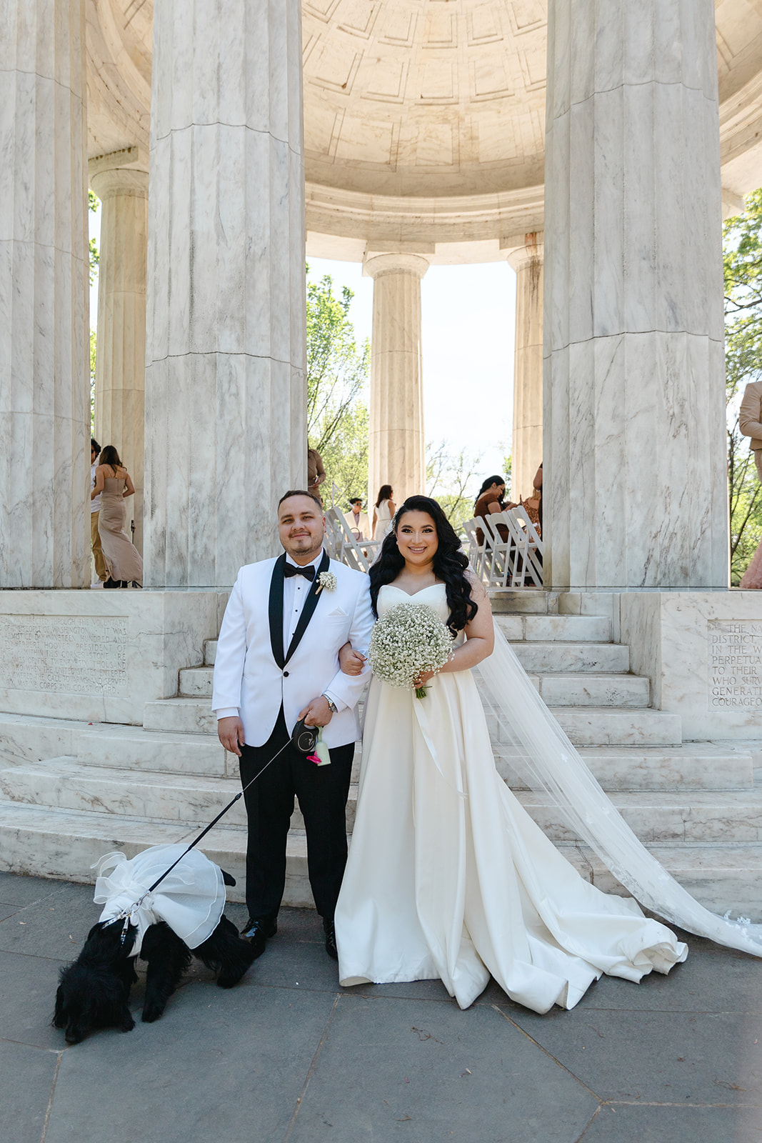 Bride and groom standing at the Washington, DC War Memorial after their ceremony, wearing classic formal attire with their dog dressed for the wedding.