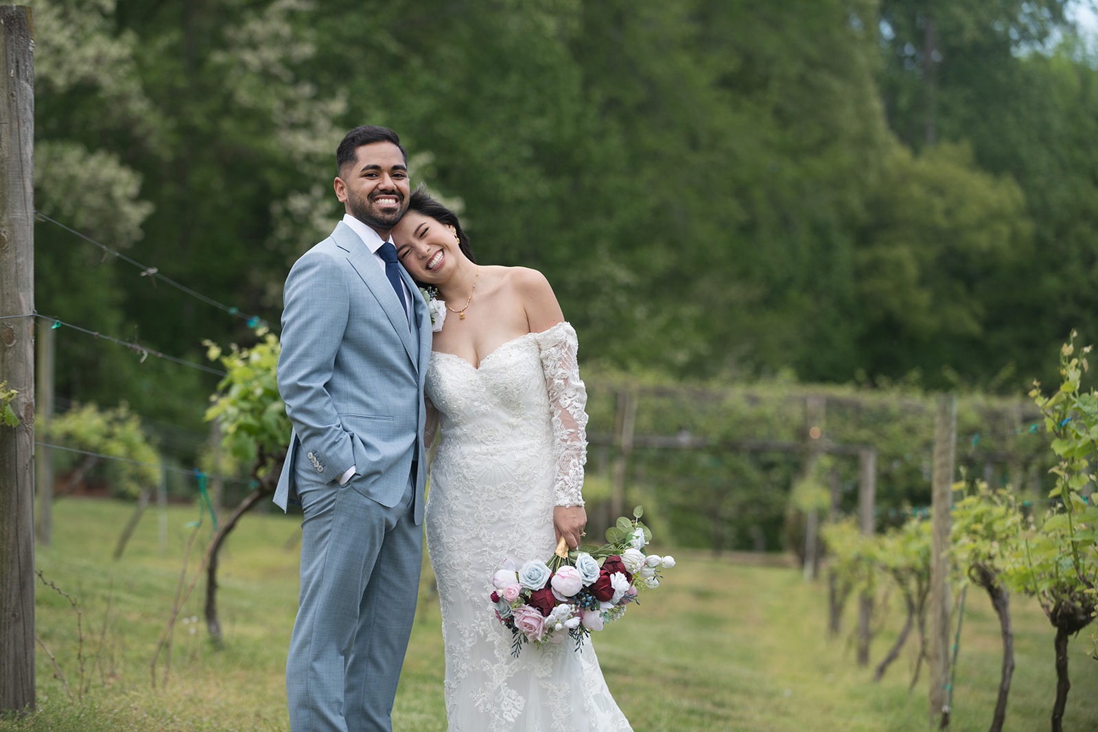 couple in a vineyard field leaning into one another with wide holding bouquet down