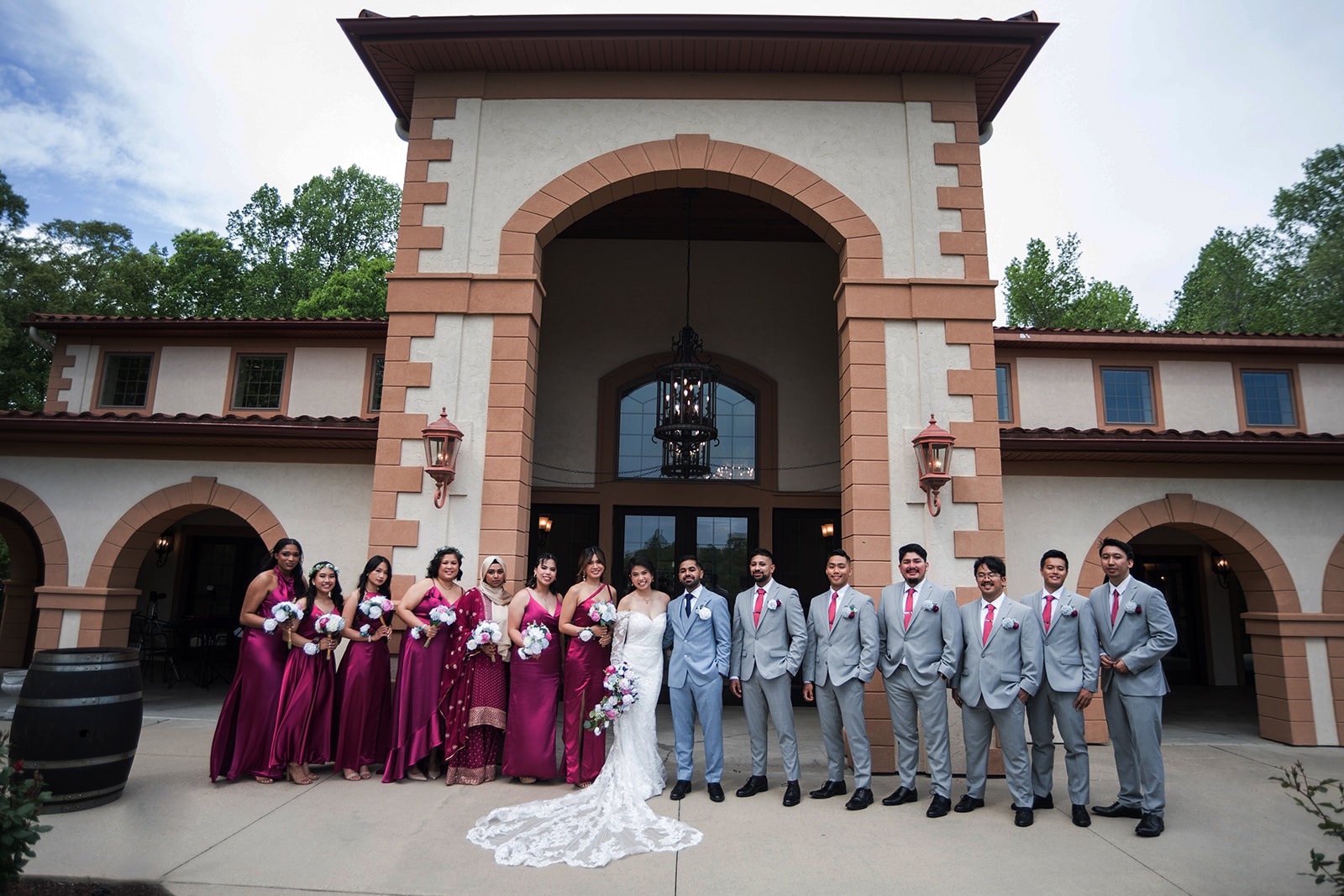 wedding part photo in from of arch way with groom to the right in a light blue suit and groomsmen wearing grey suits with pink ties. The bride in a white dress to the left with bridesmaids holding bouquets and in magenta dresses