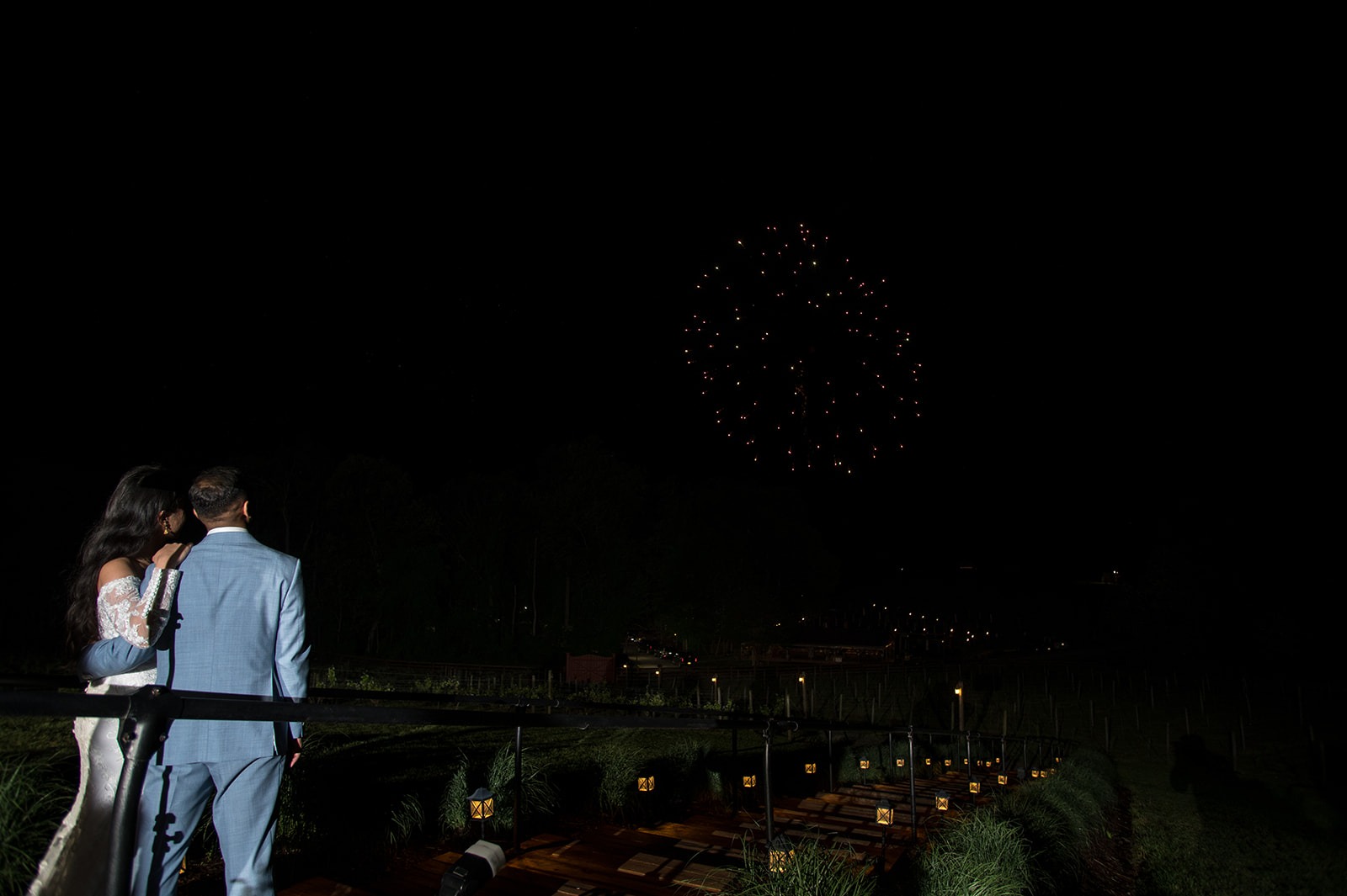 bride and groom holding each other while watching fireworks on a small wooden bridge